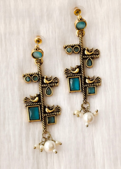 A pair of bohemian-style gold-plated dangle earrings with turquoise blue stone inlays, small bird motifs, and dangling white pearl beads, displayed on a light wood background.