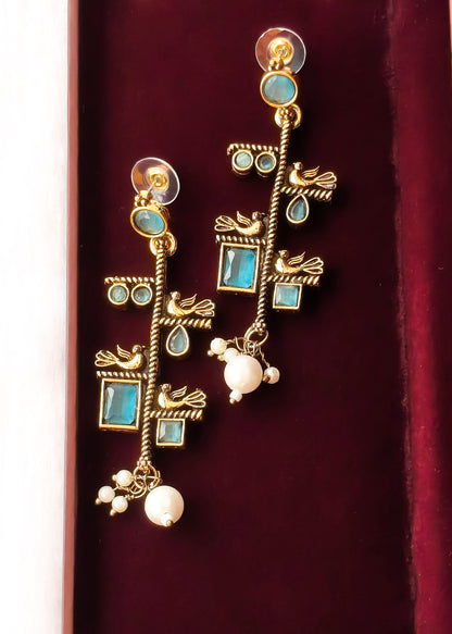 A pair of gold dangle earrings with turquoise blue stones, small bird motifs, and white pearl-like beads, displayed on a deep burgundy velvet surface.