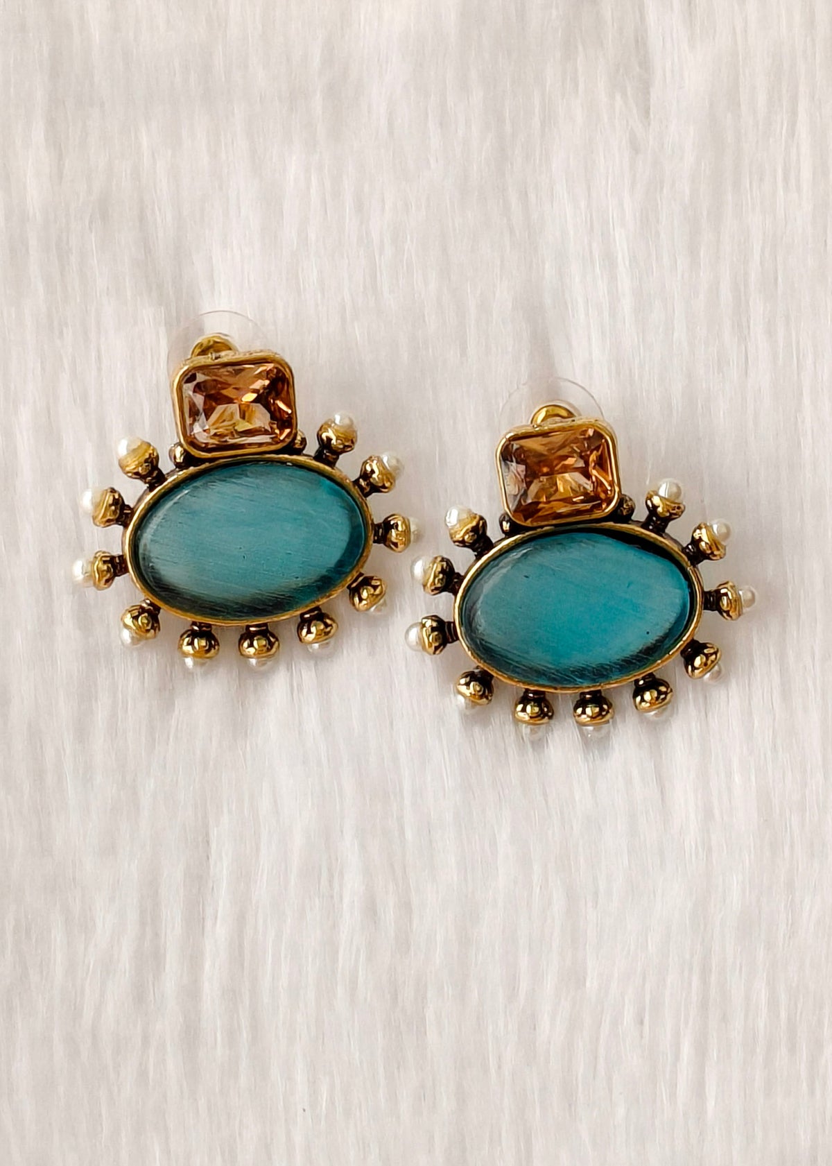 A pair of gold-toned statement earrings featuring oval turquoise-blue stone centers, a small square gold stone above, and a border of tiny pearl-like beads and gold beads, displayed on a white textured background.