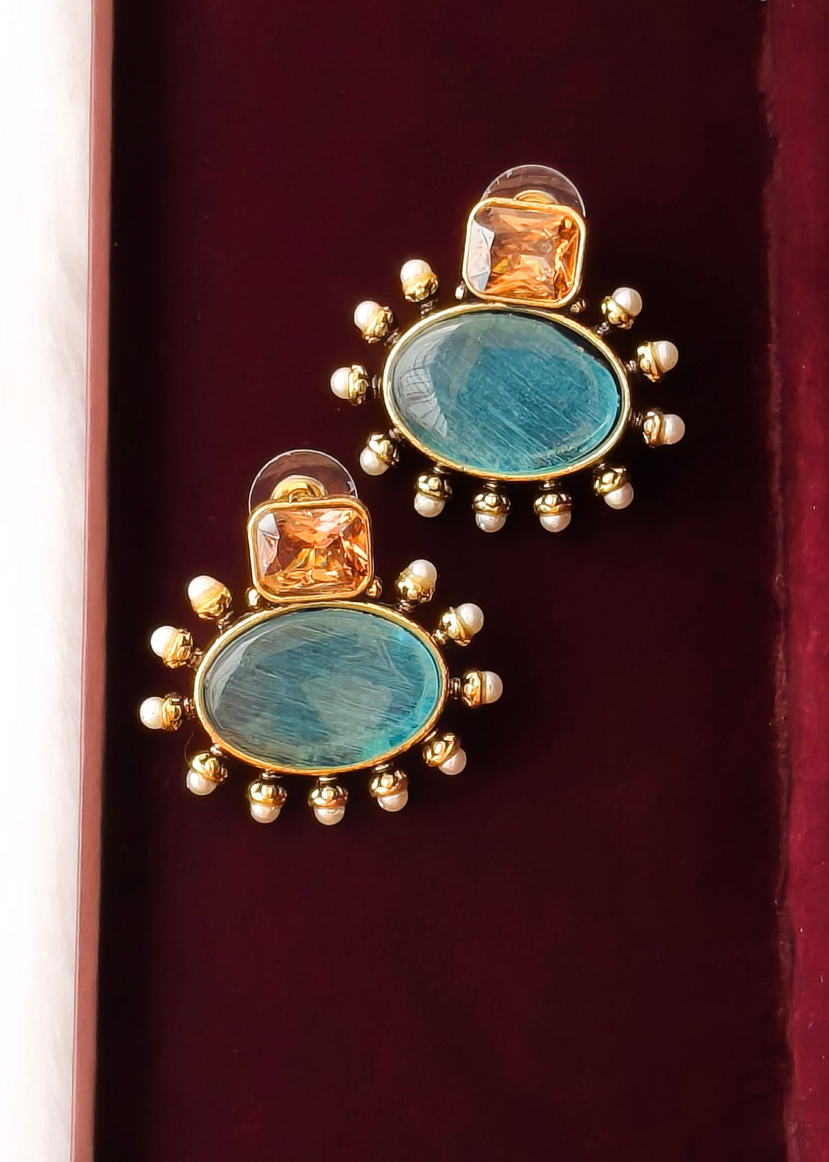 A pair of gold-plated statement earrings featuring oval turquoise-blue stones surrounded by small gold beads, with a smaller square amber-orange stone above, displayed on a dark red velvet surface.