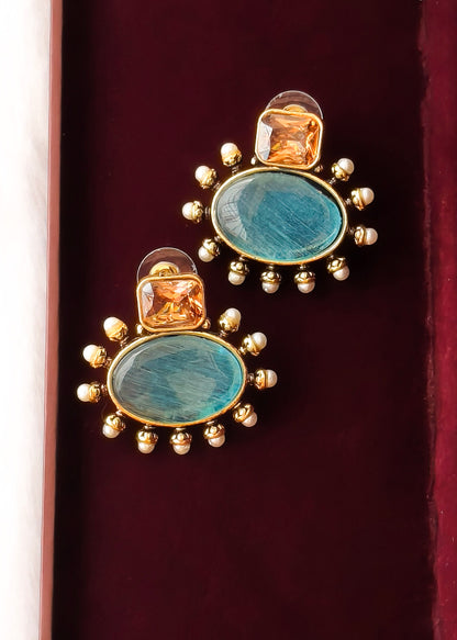 A pair of gold-plated statement earrings featuring oval turquoise-blue stones surrounded by small gold beads, with a smaller square amber-orange stone above, displayed on a dark red velvet surface.