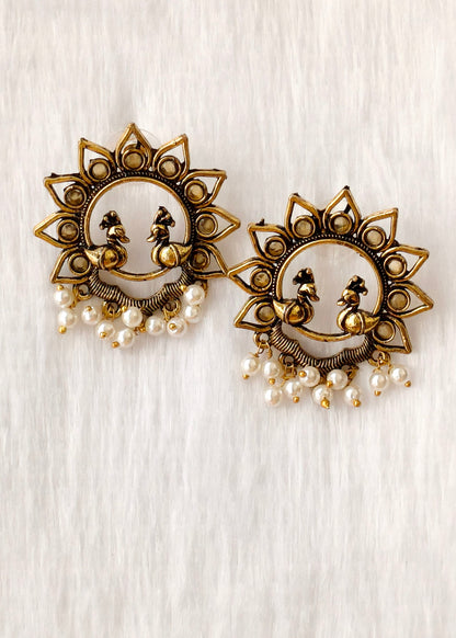 A pair of ornate gold-toned statement earrings featuring a sunburst design, two peacock motifs, and dangling pearl-like beads, displayed on a light textured fabric background.