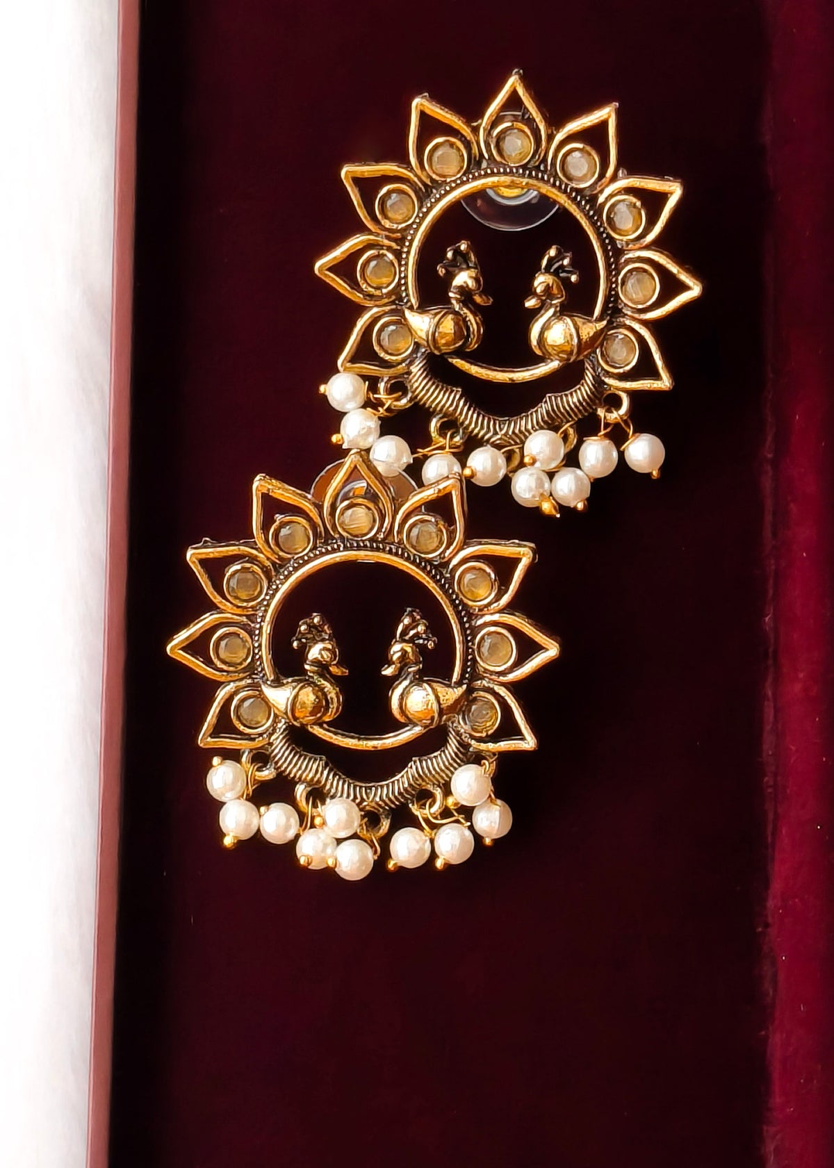 A pair of ornate gold-toned earrings with sunburst motifs, peacock details, and dangling pearl beads, displayed on a dark burgundy velvet surface.