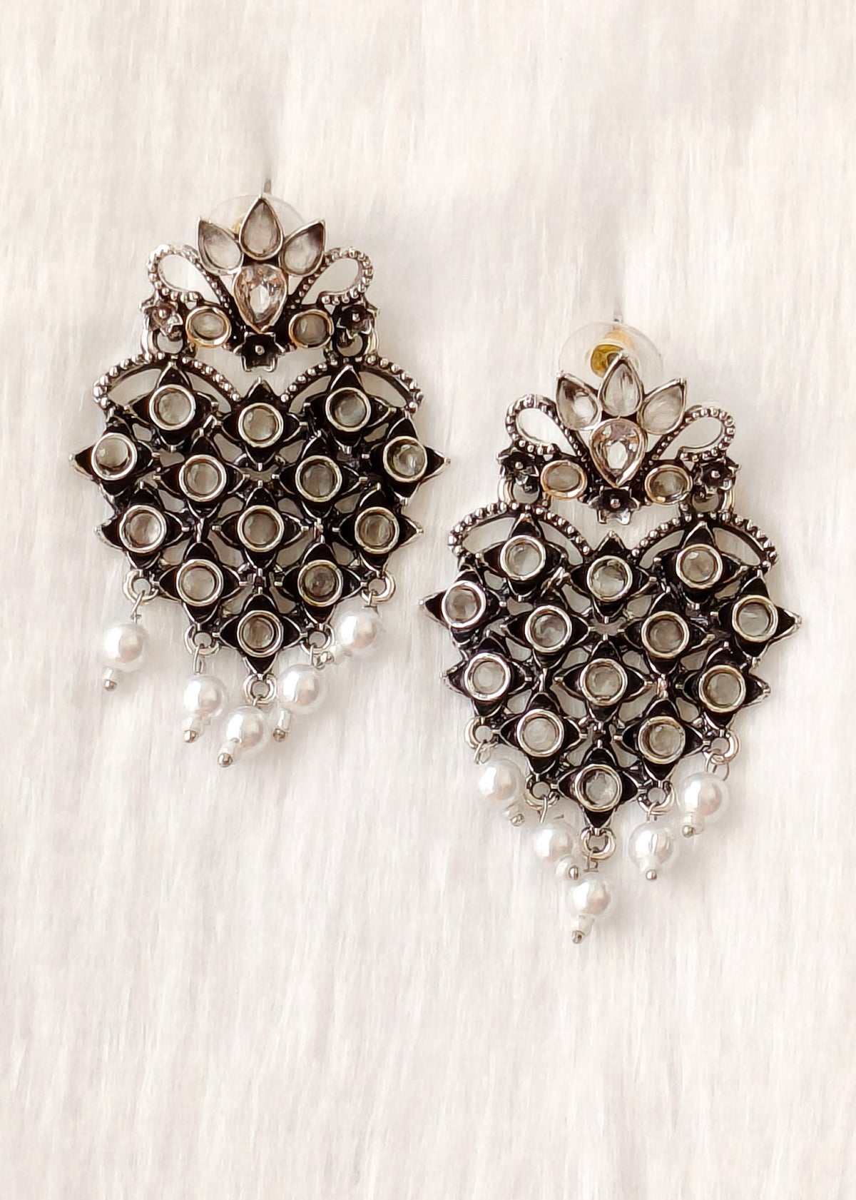 A pair of ornate silver-toned statement earrings with intricate filigree, clear stone accents, and dangling white faux pearls, displayed on a white textured fabric background.