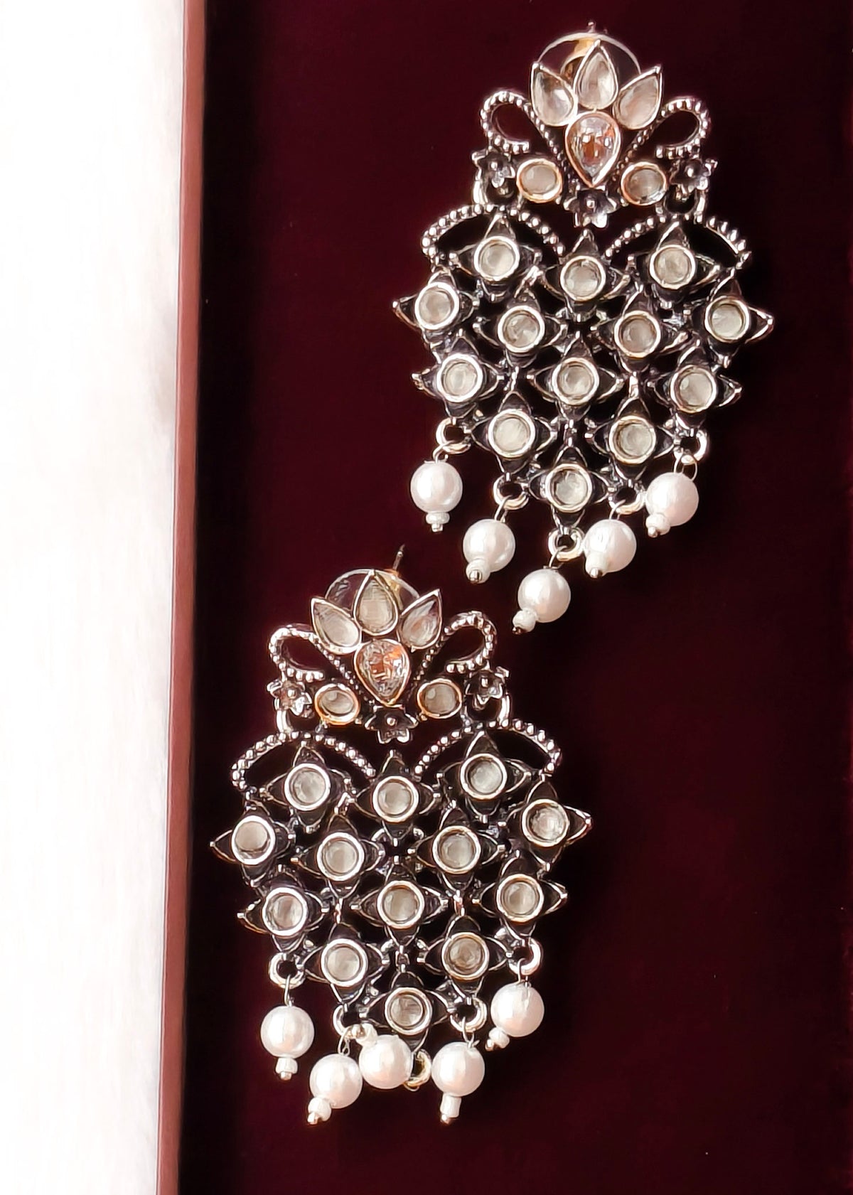 A pair of ornate silver-toned chandelier earrings with white pearl drops and clear rhinestone embellishments, displayed on a dark red velvet surface.