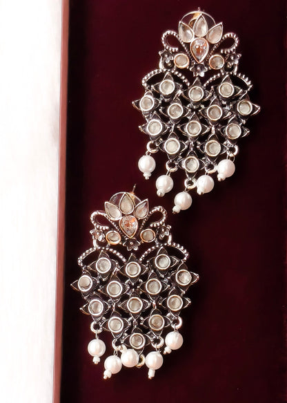 A pair of ornate silver-toned chandelier earrings with white pearl drops and clear rhinestone embellishments, displayed on a dark red velvet surface.