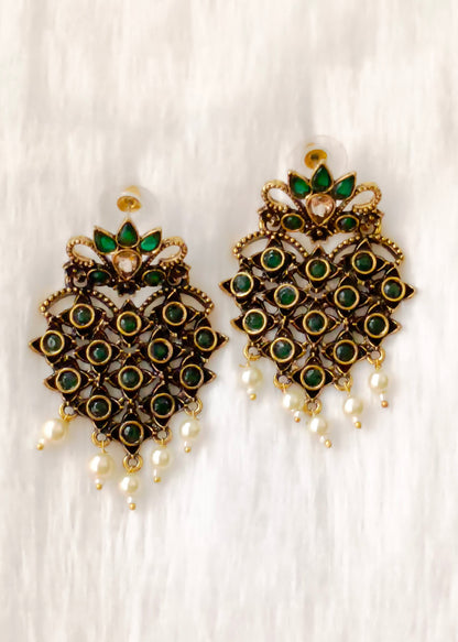 A pair of ornate gold-toned traditional Indian earrings decorated with green stones, a central clear stone, and dangling white pearl beads, displayed on a white textured fabric background.