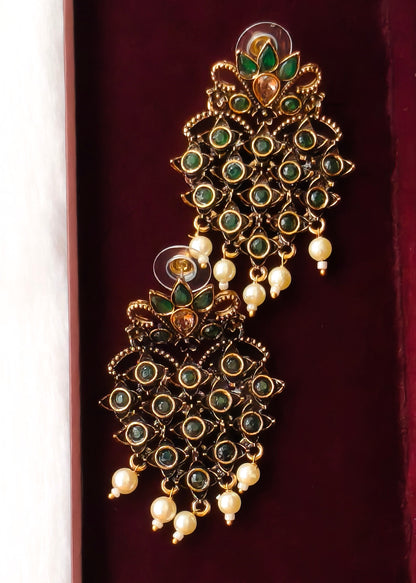 A pair of ornate gold-plated Indian chandelier earrings decorated with green enamel, green stones, and white pearl-like drops, displayed on a dark red velvet surface.
