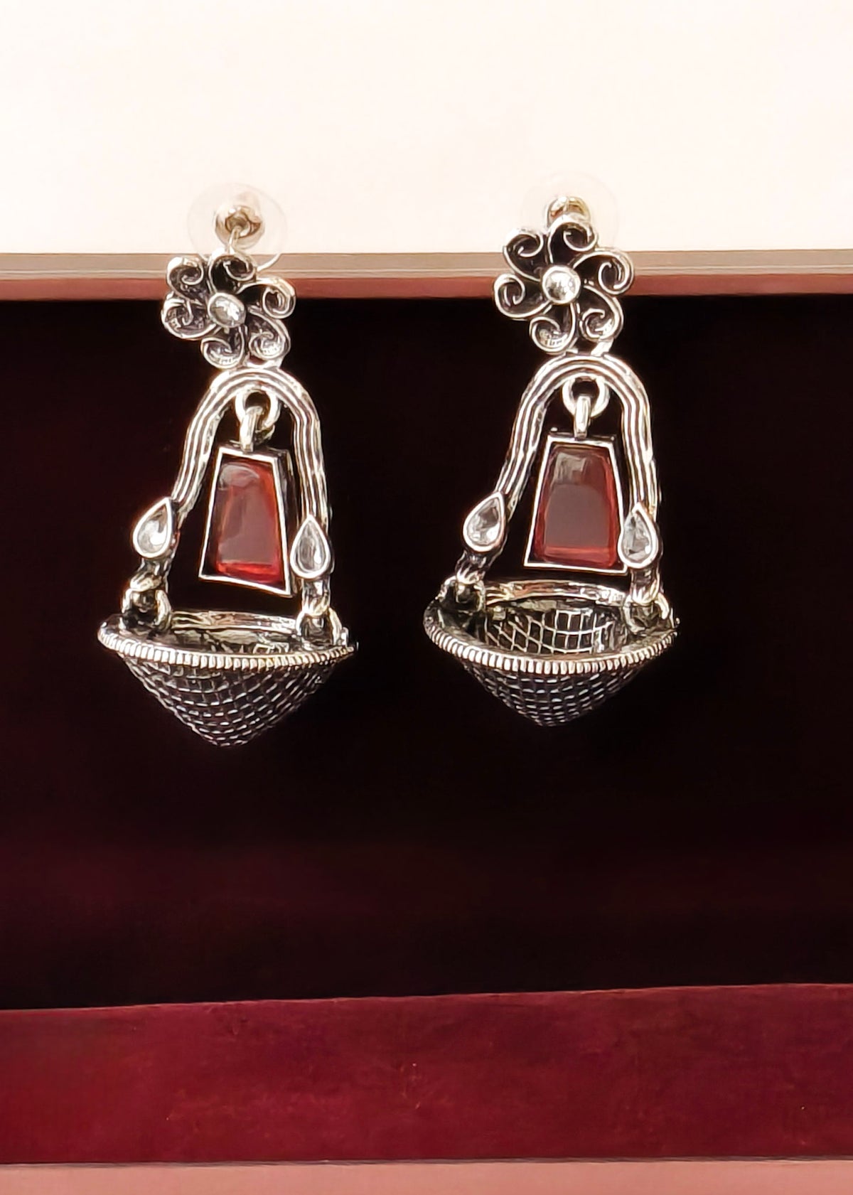 A pair of silver-toned dangle earrings with red rectangular stone accents, displayed inside a dark red velvet jewelry box.