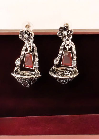 A pair of silver-toned dangle earrings with red rectangular stone accents, displayed inside a dark red velvet jewelry box.