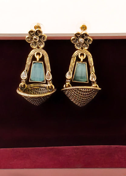 A pair of ornate gold-toned dangle earrings with rectangular light blue-green stone centers and small clear stones, displayed against a dark burgundy velvet surface.