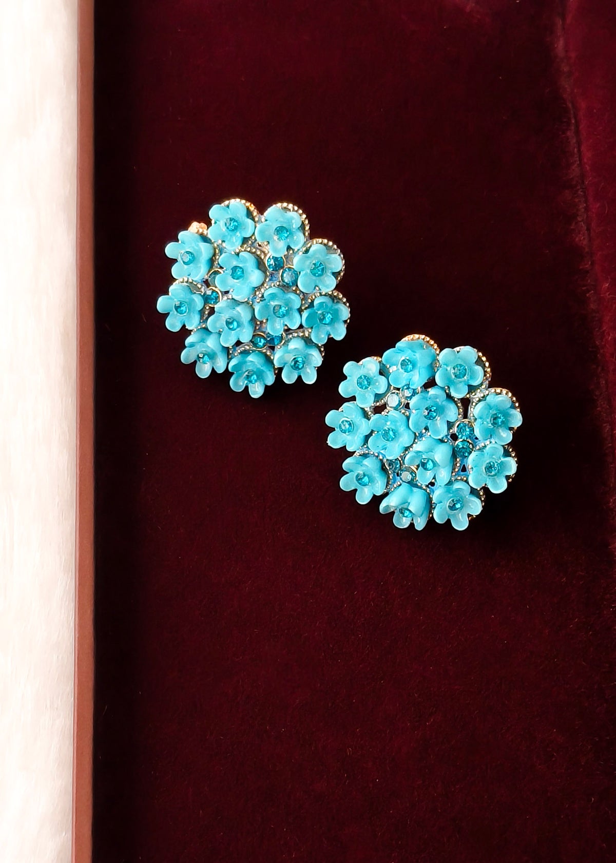 A pair of turquoise blue flower-shaped stud earrings with gold-tone detailing, displayed on a deep burgundy velvet surface.
