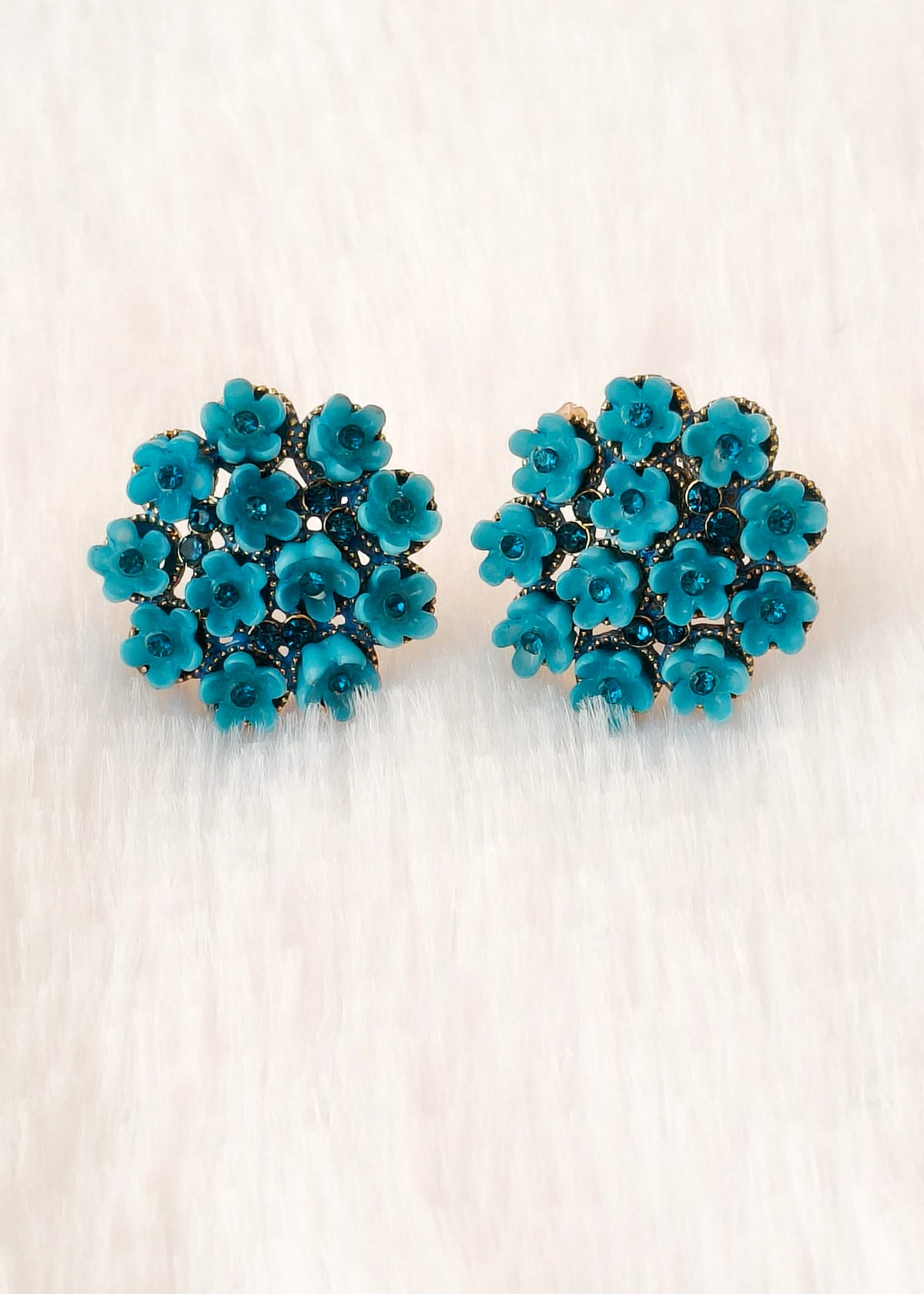 A pair of teal blue flower-shaped stud earrings with gold-tone metal detailing, displayed on a soft, light beige textured surface.