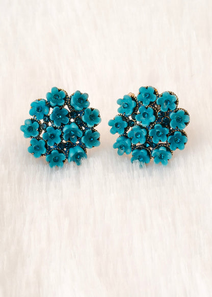 A pair of teal blue flower-shaped stud earrings with gold-tone metal detailing, displayed on a soft, light beige textured surface.
