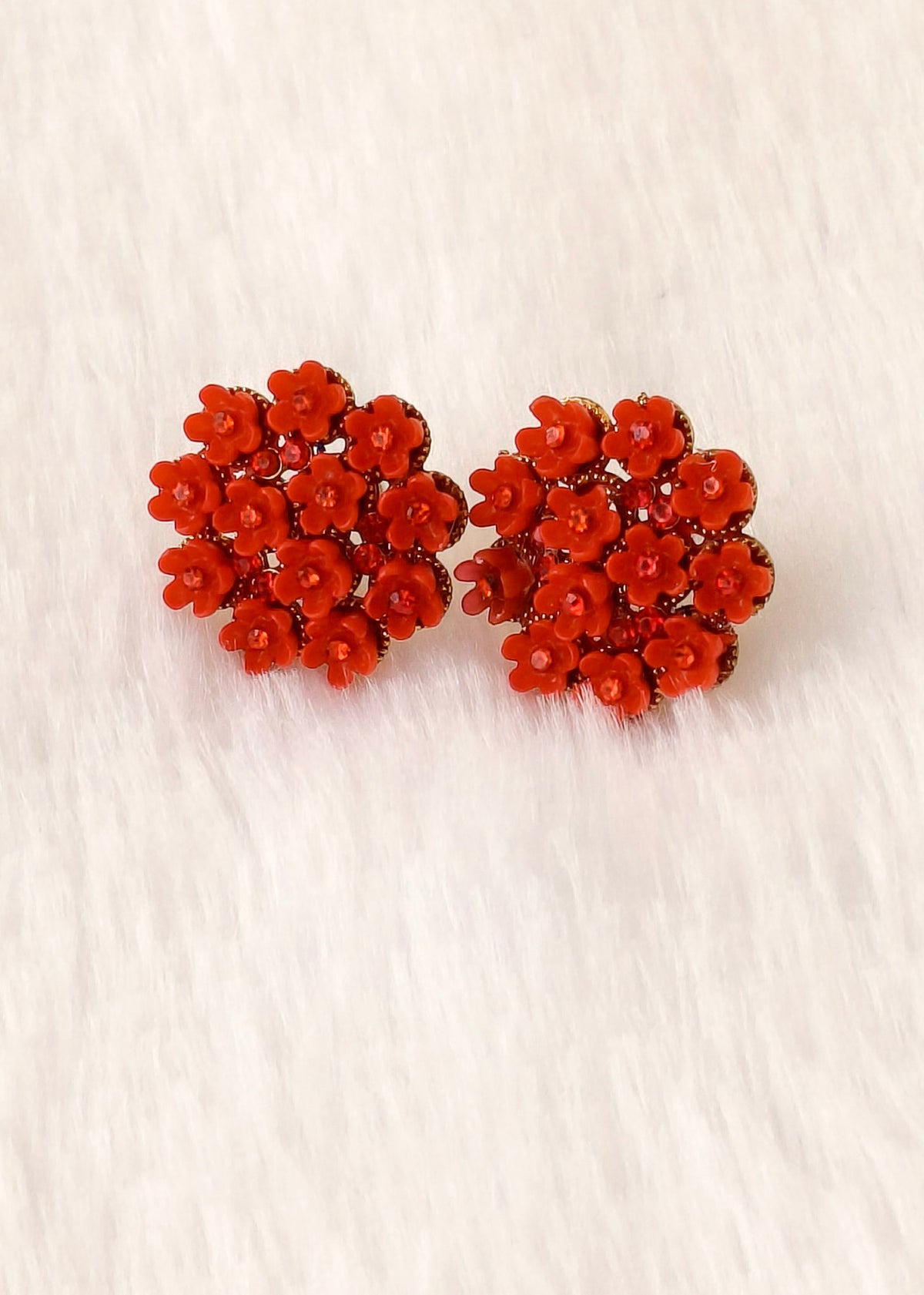 A pair of red floral stud earrings with small faceted clear stones, displayed on a white faux fur surface.
