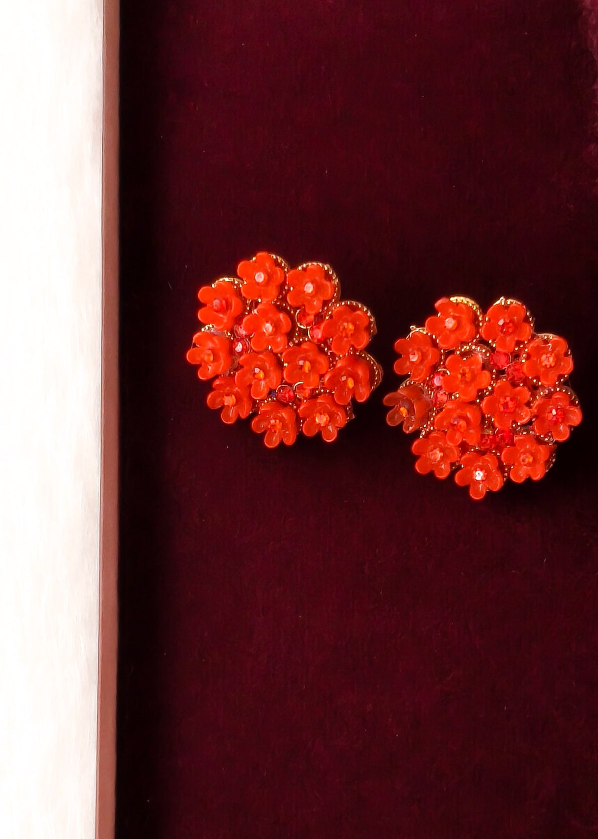 A pair of bright orange-red floral stud earrings with gold-tone metal backing, displayed on a dark burgundy velvet surface next to a light wood edge.