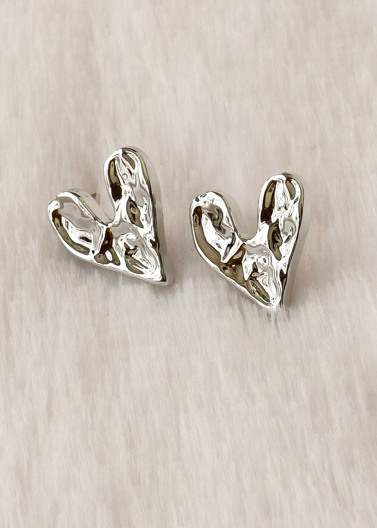 A pair of textured silver heart-shaped stud earrings on a light beige fabric background.