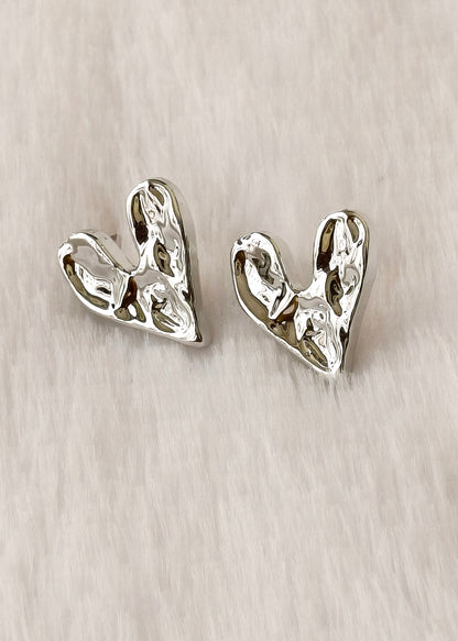A pair of textured silver heart-shaped stud earrings on a light beige fabric background.