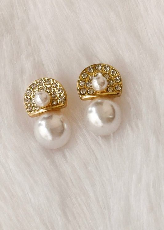 A pair of gold and pearl stud earrings with rhinestone-embellished semicircles and large white pearls, displayed on a soft white faux fur background.