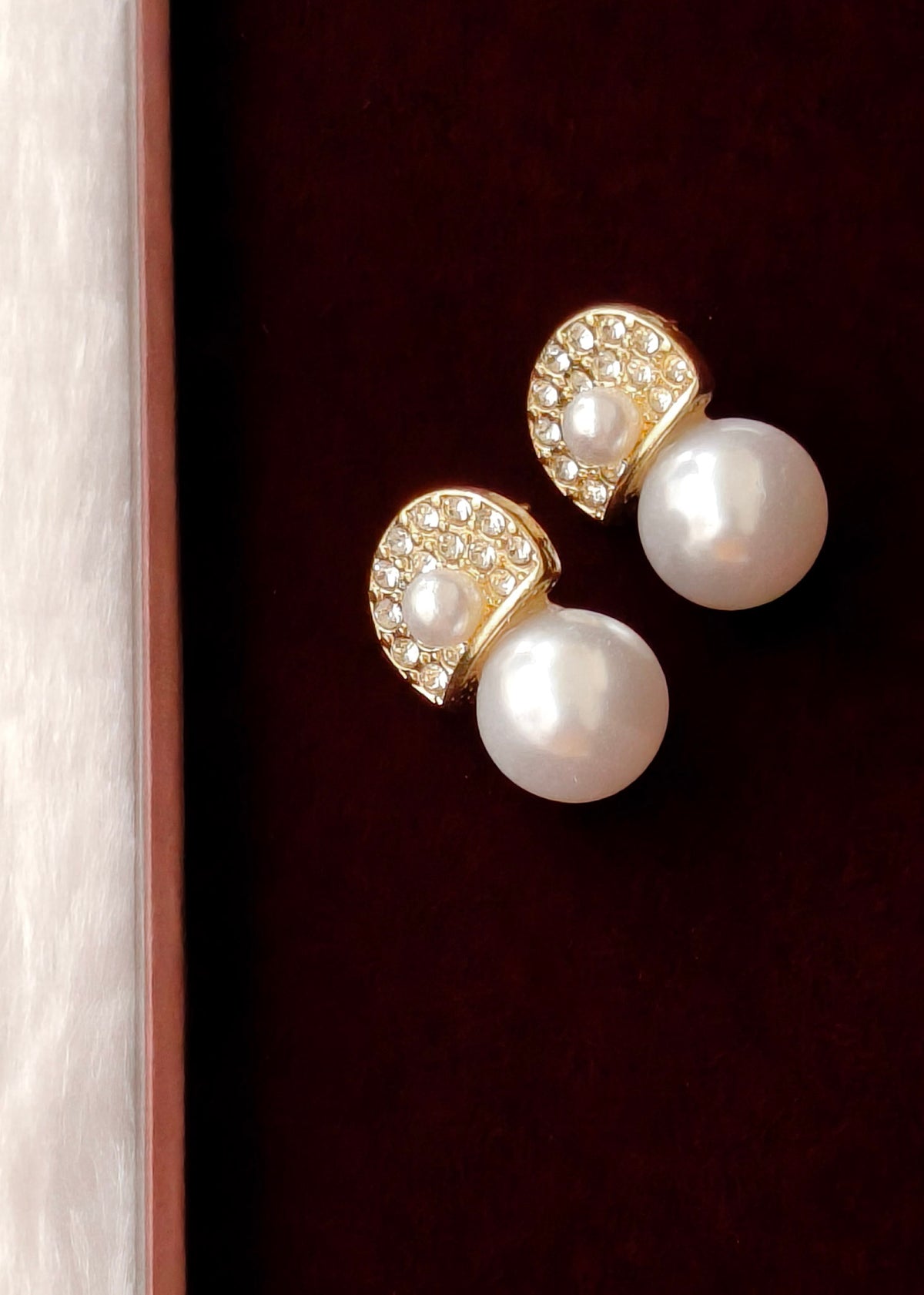 A pair of gold and pearl stud earrings displayed on a dark burgundy velvet surface next to a light wood border.