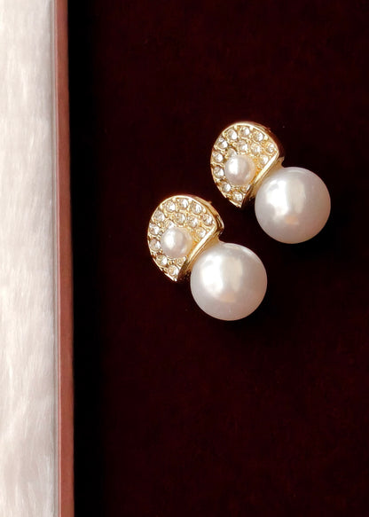 A pair of gold and pearl stud earrings displayed on a dark burgundy velvet surface next to a light wood border.