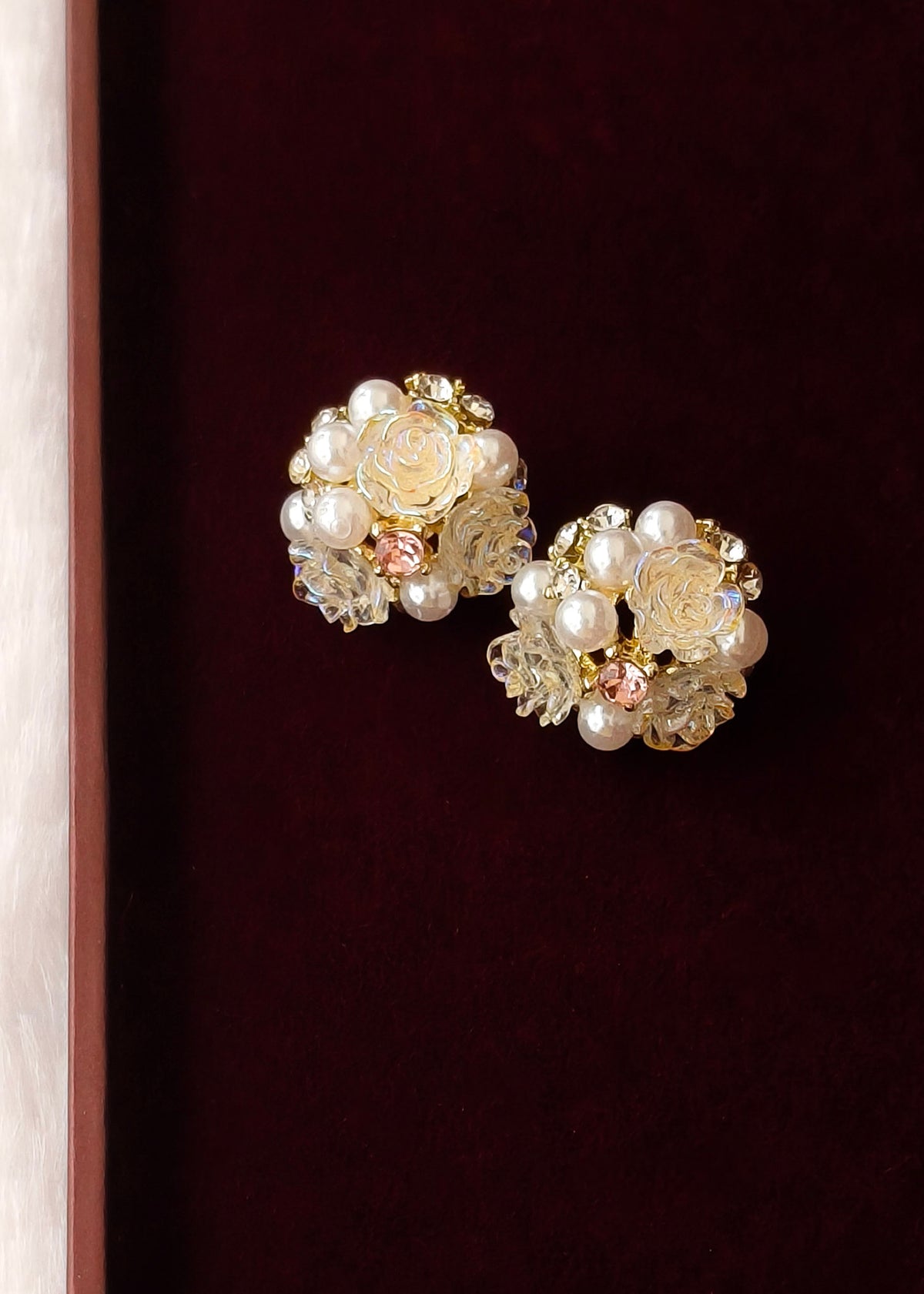 A pair of vintage-style floral stud earrings featuring cream-colored faux pearls, clear faceted crystals, and small gold-toned rose details, displayed on a dark burgundy velvet surface.