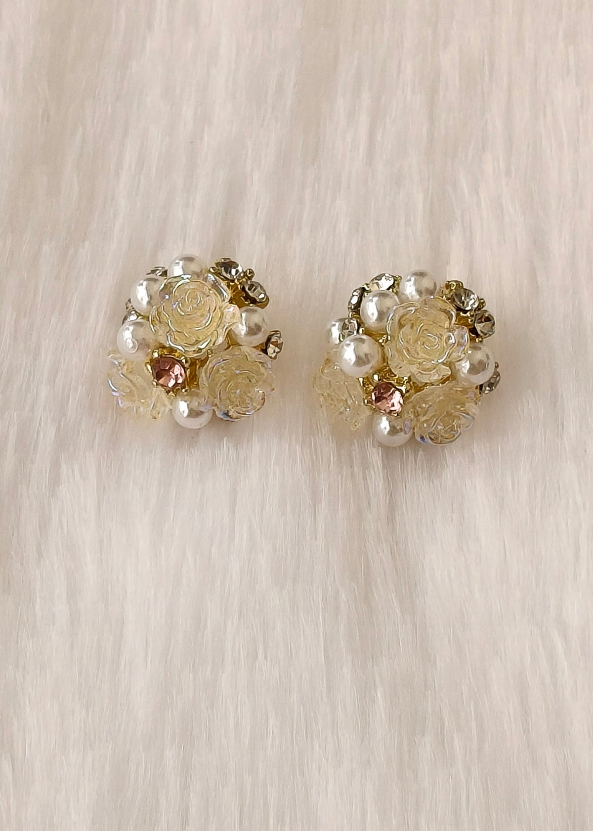 A pair of vintage-style floral stud earrings featuring cream-colored faux roses, white faux pearls, and small gold-toned metal details, displayed on a soft beige faux fur background.
