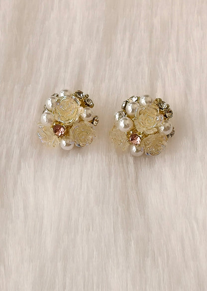 A pair of vintage-style floral stud earrings featuring cream-colored faux roses, white faux pearls, and small gold-toned metal details, displayed on a soft beige faux fur background.