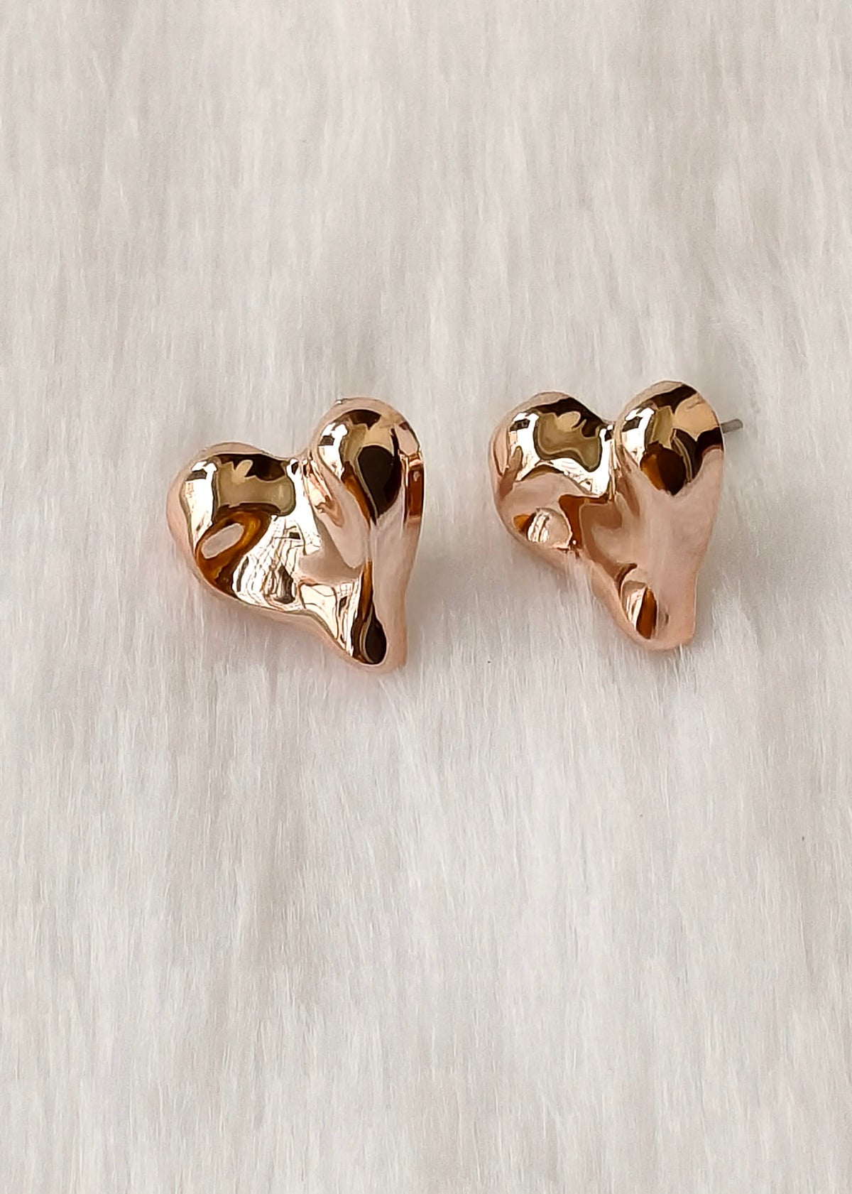 A pair of rose gold heart-shaped stud earrings with a polished, organic wavy texture, displayed on a soft white faux fur background.