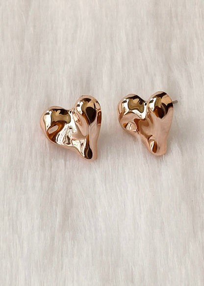 A pair of rose gold heart-shaped stud earrings with a polished, organic wavy texture, displayed on a soft white faux fur background.
