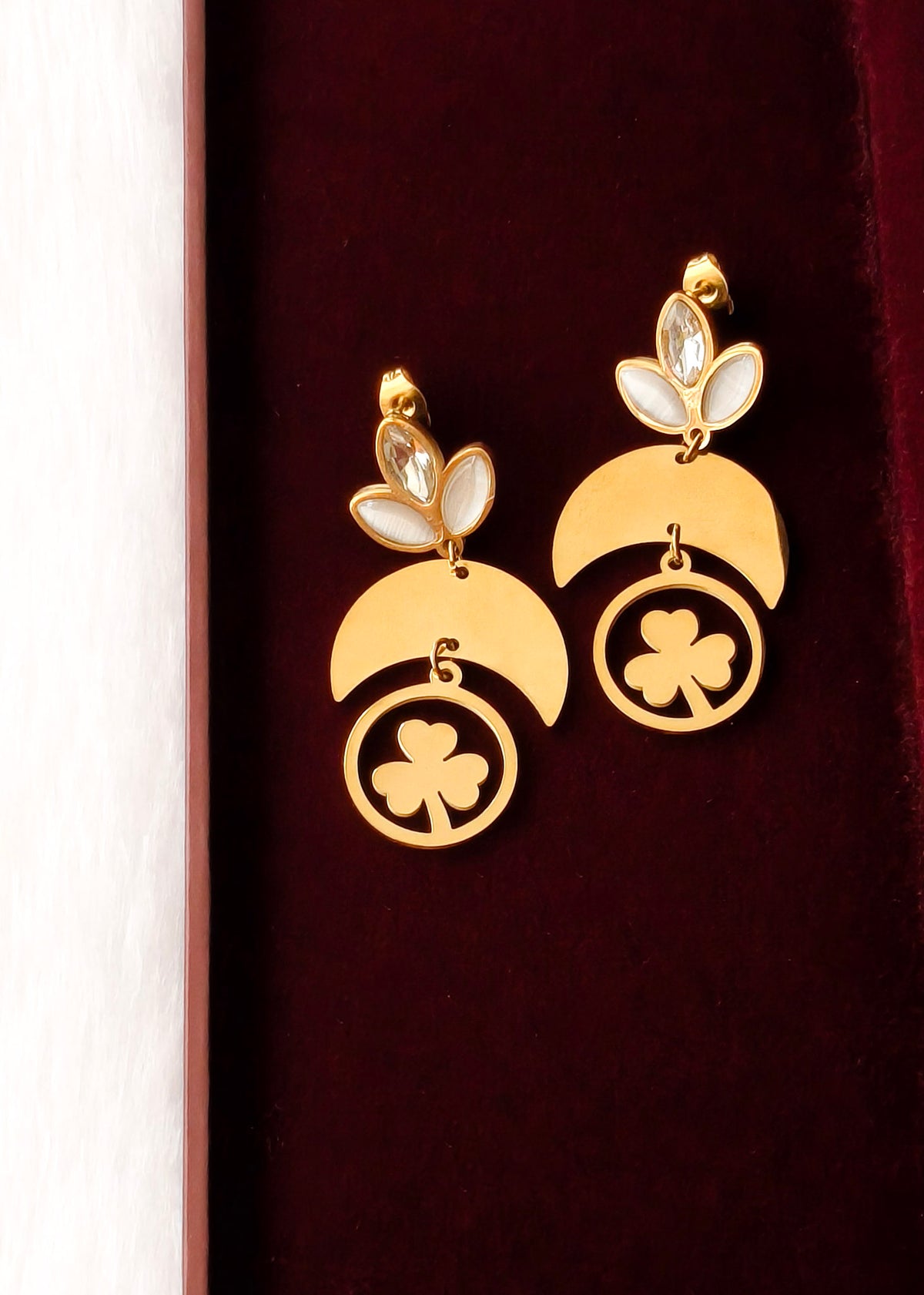 A pair of gold dangle earrings with white pearl-like accents and shamrock motifs, displayed on a dark red velvet surface.