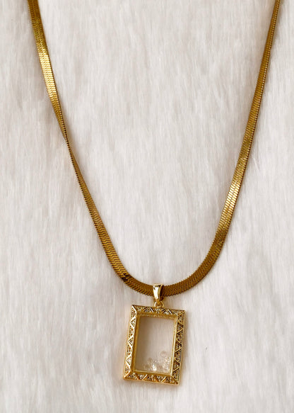A gold snake chain necklace with a rectangular gold pendant frame, displayed on a white faux fur background.