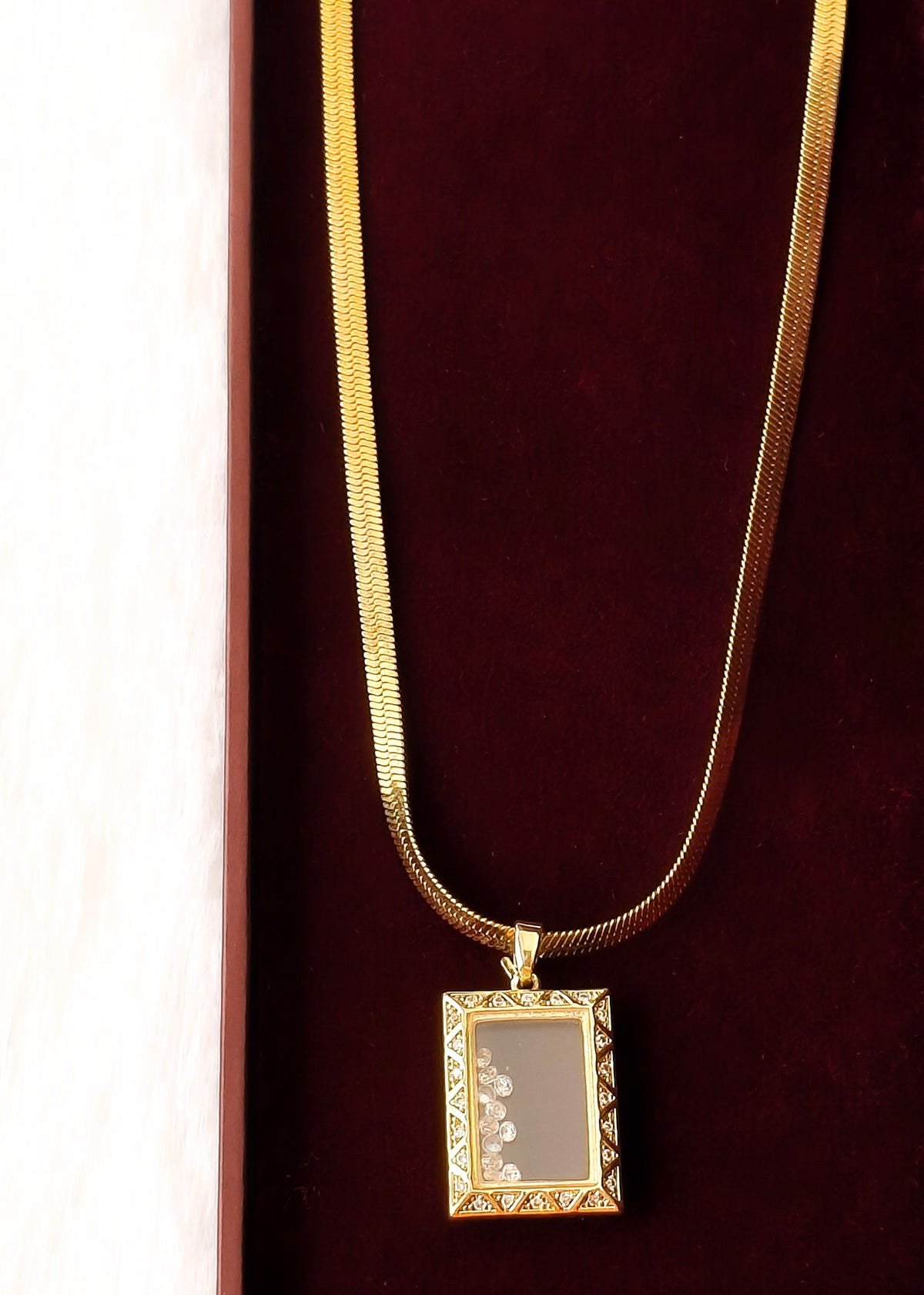 A gold chain necklace with a rectangular pendant featuring a floral design on a dark gray background, displayed on a dark red velvet surface.