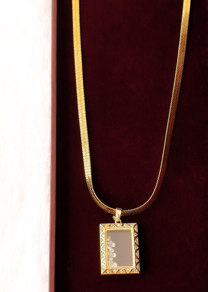A gold chain necklace with a rectangular pendant featuring a floral design on a dark gray background, displayed on a dark red velvet surface.