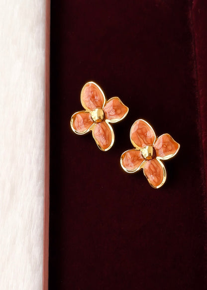 A pair of gold-tone flower-shaped stud earrings with textured orange-pink enamel petals, displayed on a dark burgundy velvet surface next to a light wood edge.