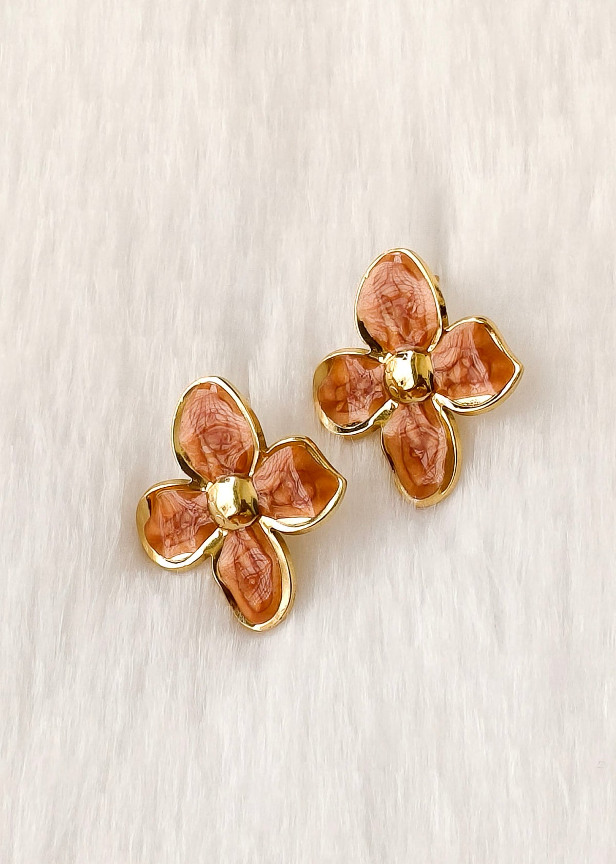 A pair of gold flower-shaped stud earrings with textured, warm brown petal inlays, displayed on a light beige textured fabric background.