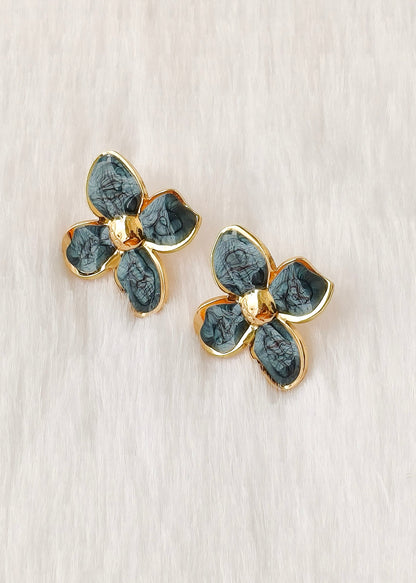 A pair of gold-tone flower-shaped earrings with textured dark green enamel petals, displayed on a light beige textured surface.