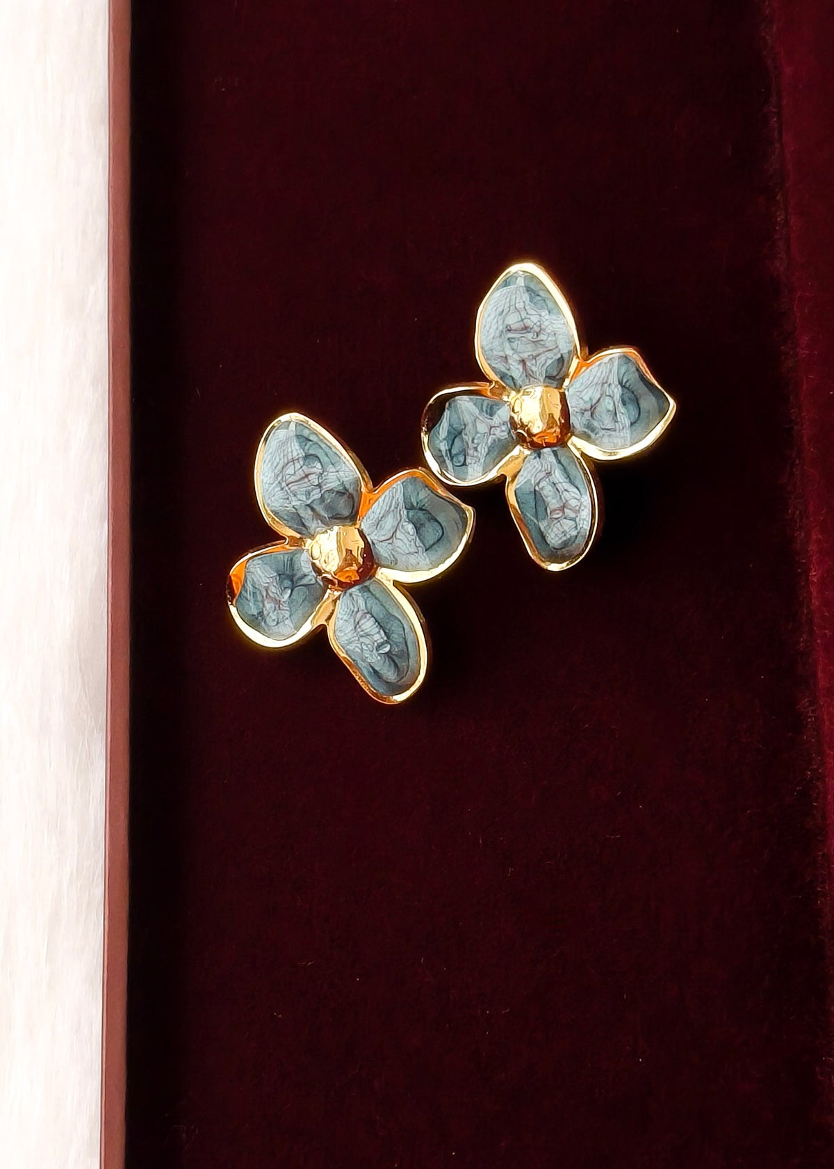 A pair of gold-tone flower-shaped earrings with light blue enamel petals, displayed on a dark red velvet surface.