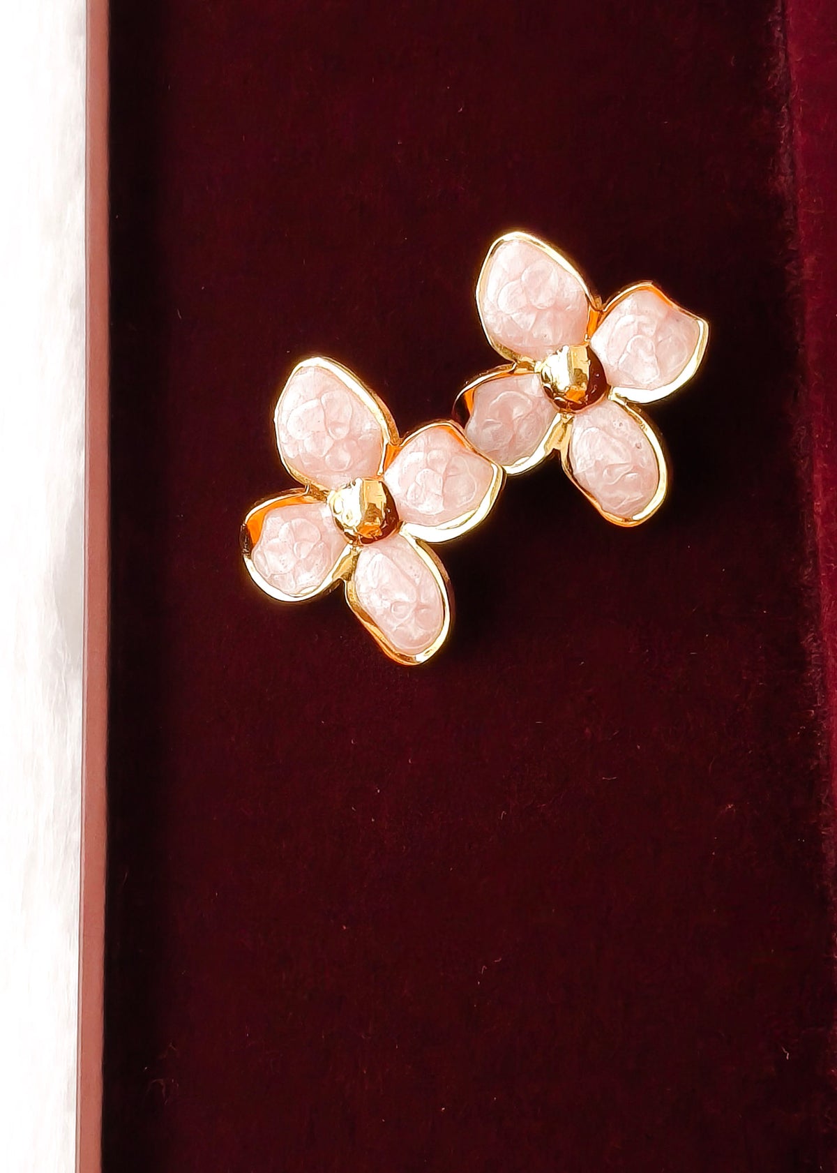 A pair of gold-tone flower-shaped earrings with pale pink stone petals, displayed on a dark burgundy velvet surface.