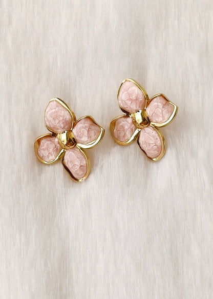 A pair of gold flower-shaped stud earrings with textured light pink petals, displayed on a soft, light beige textured fabric background.