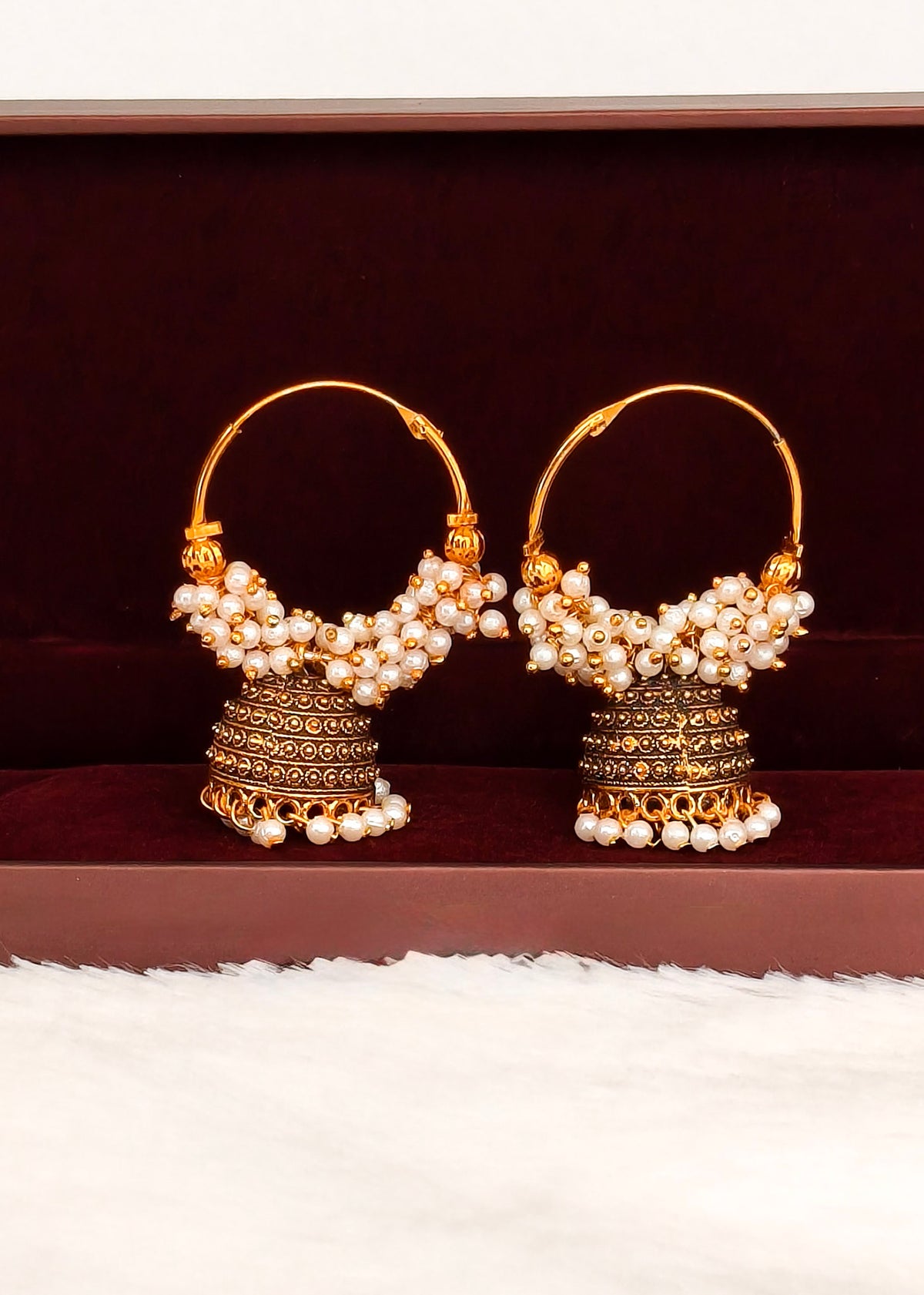 A pair of traditional Indian gold jhumka earrings adorned with white pearls, displayed on a dark brown velvet jewelry box.