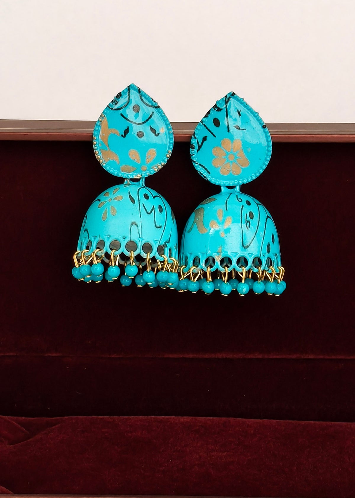 A pair of turquoise blue jhumka earrings with gold floral engravings and dangling bead tassels, displayed on a dark red velvet surface against a light beige background.