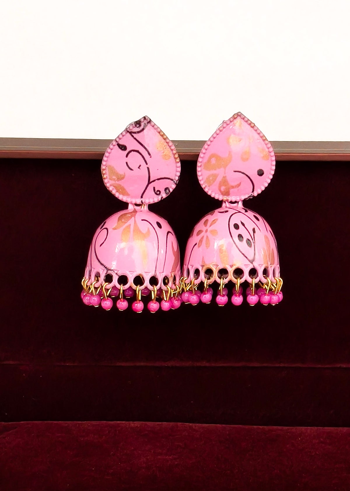 A pair of pink jhumka earrings with gold and black painted patterns, hanging from gold-toned chains with dangling pink beads, displayed against a dark red velvet surface.