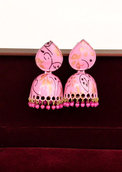 A pair of pink jhumka earrings with gold and black painted patterns, hanging from gold-toned chains with dangling pink beads, displayed against a dark red velvet surface.