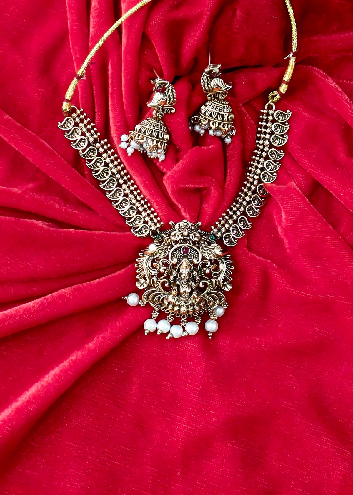 An ornate Indian jewelry set featuring a necklace and matching earrings with detailed silver-toned filigree, white pearl beads, and red stone accents, displayed on a draped red velvet fabric background.