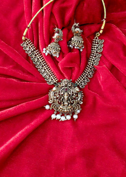 An ornate Indian jewelry set featuring a necklace and matching earrings with detailed silver-toned filigree, white pearl beads, and red stone accents, displayed on a draped red velvet fabric background.