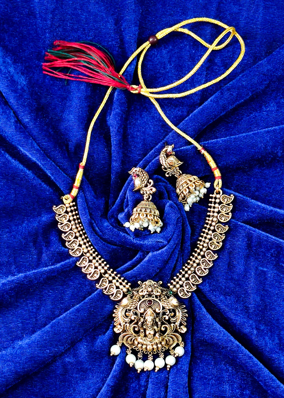 An ornate gold-toned traditional necklace set with matching earrings, featuring a large central pendant, beaded necklaces, and red tassels, arranged on a deep blue velvet background.