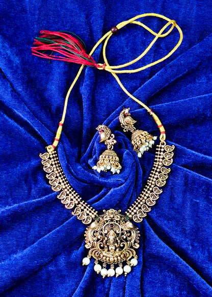 An ornate gold-toned traditional necklace set with matching earrings, featuring a large central pendant, beaded necklaces, and red tassels, arranged on a deep blue velvet background.