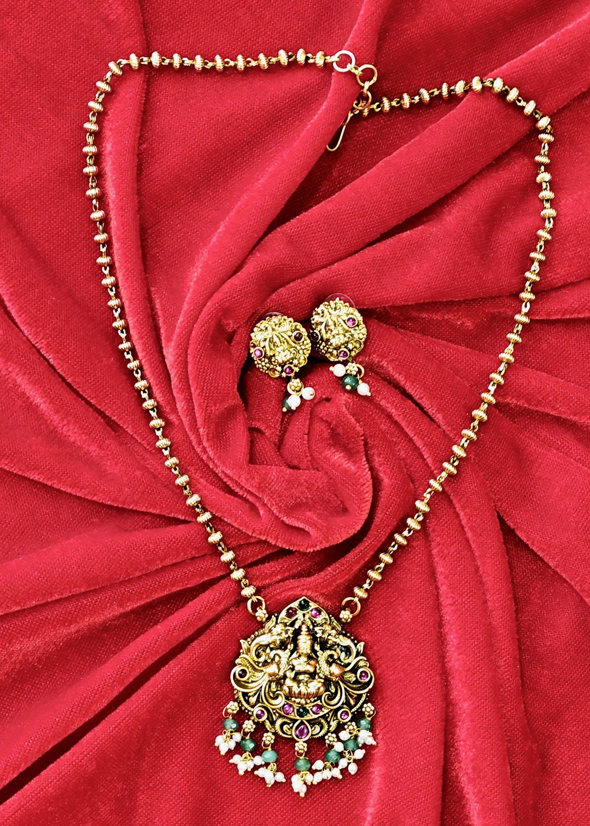 Gold-toned traditional necklace and matching earrings with ornate filigree detailing and multicolored gemstone accents, displayed on a red fabric background.