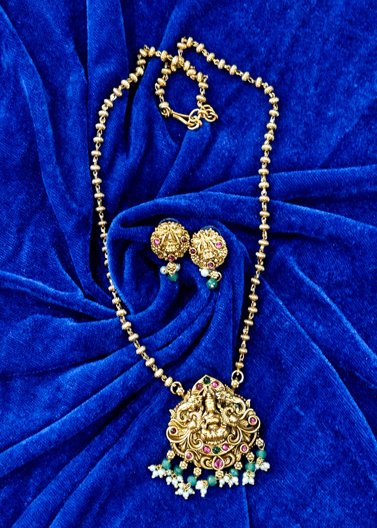 Gold-toned traditional necklace and matching earrings displayed on a rich royal blue velvet fabric background. The necklace features an ornate Lakshmi pendant embellished with small pink, green, and white stones, and the matching earrings have similar floral gold tops with blue and green bead drops.